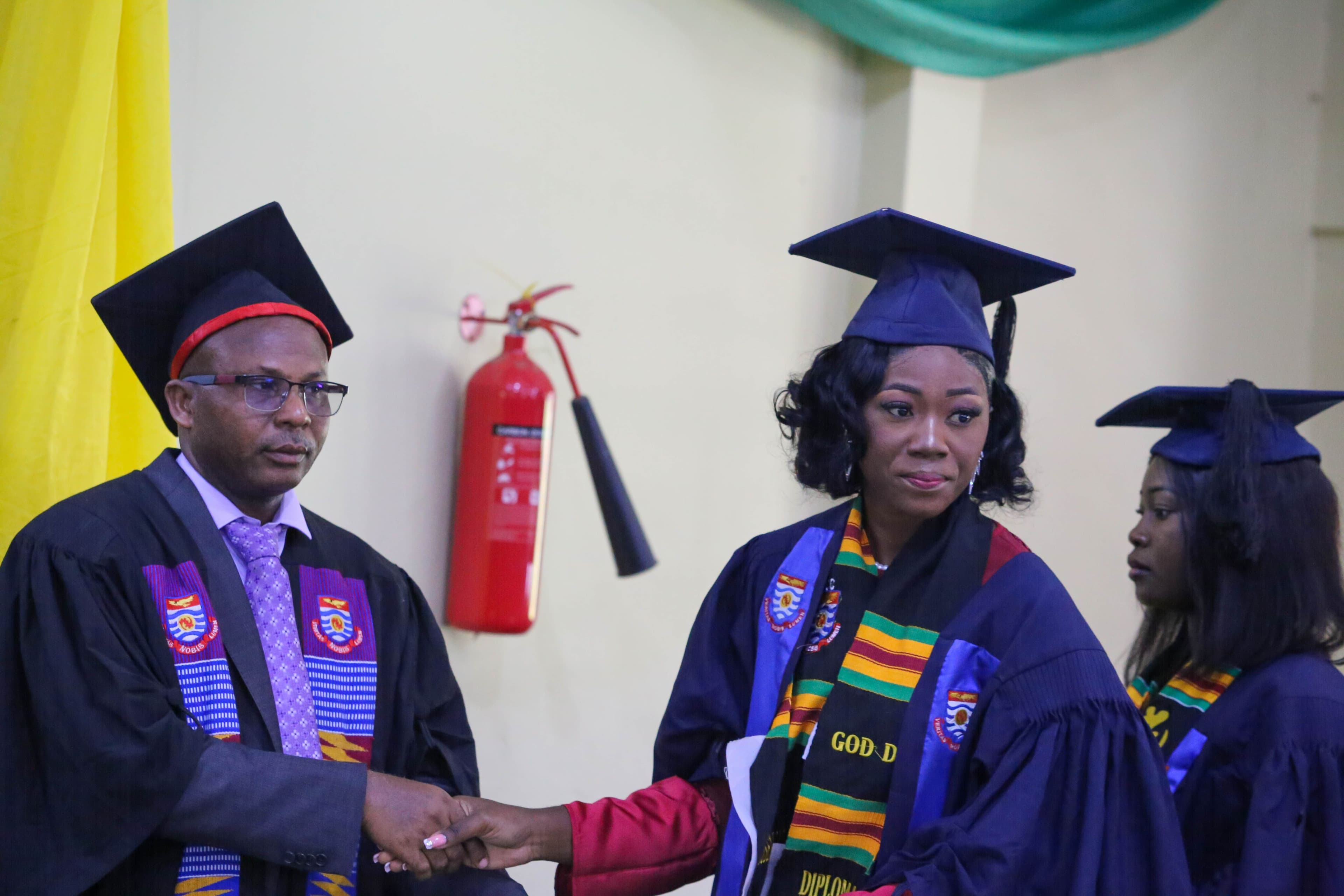 A male graduate in black robes shakes hands with a female graduate in blue robes.
